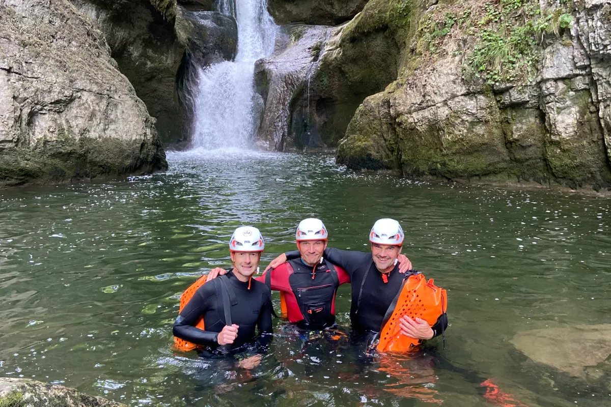 Trois participants en combinaison néoprène intégrale dans une vasque au pied d'une cascade, canyoning dans le Jura